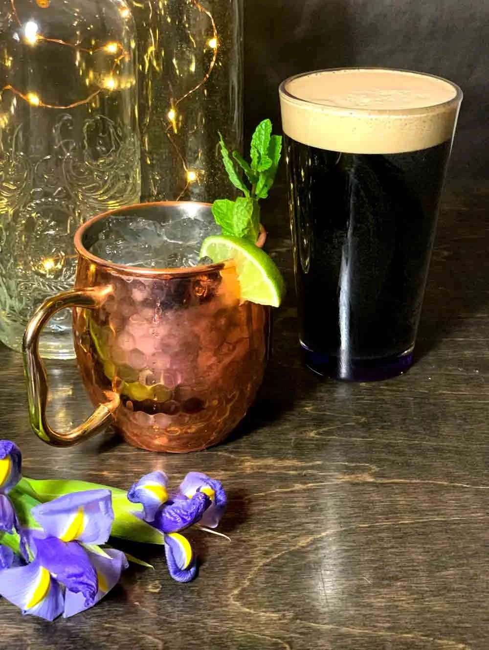 A copper mug of a mule cocktail, garnished with a lime wedge and mint, next to a pint of Guinness with a creamy head, with decorative jars and flowers on a dark wooden table.