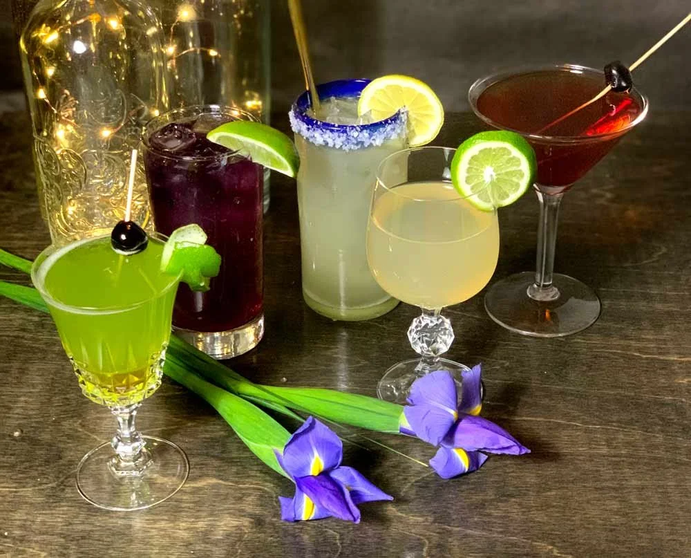 Six colorful cocktails garnished with lemon, lime, or cherry, decorated with purple irises, set on a dark wooden table with a gold ornate jar in the background.