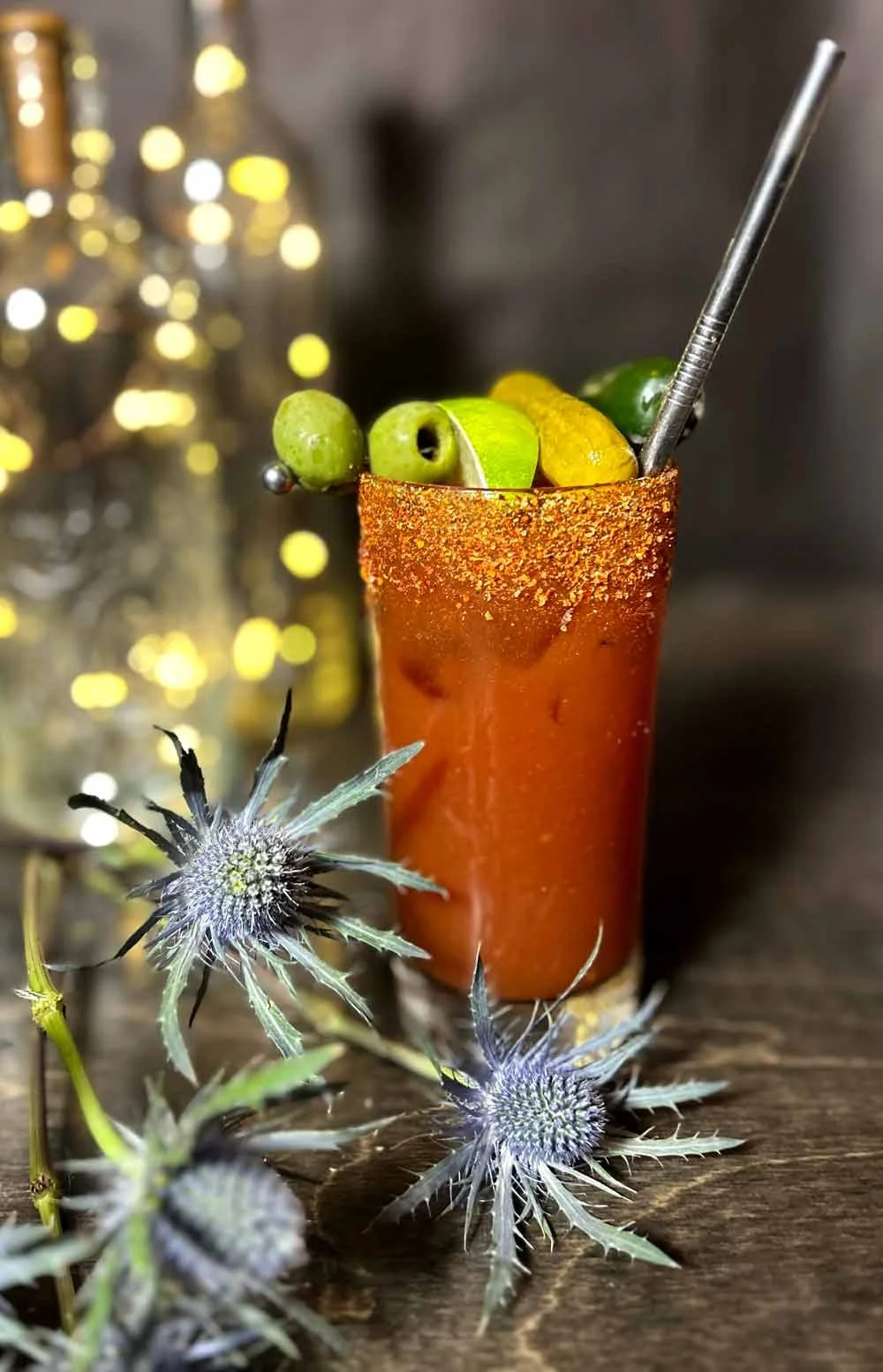 A tall glass of Bloody Mary cocktail garnished with green olives and a lime wedge, with a salted rim, on a wooden surface decorated with blue thistle flowers.