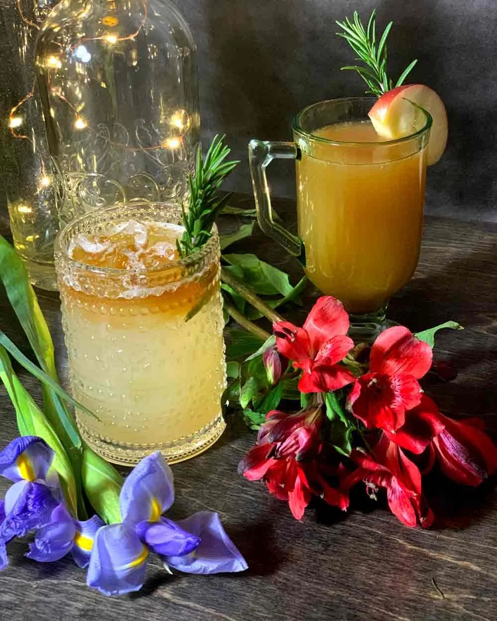 Two craft cocktails decorated with rosemary sprigs and apple slices, placed alongside colorful flowers on a wooden surface with a dark background.