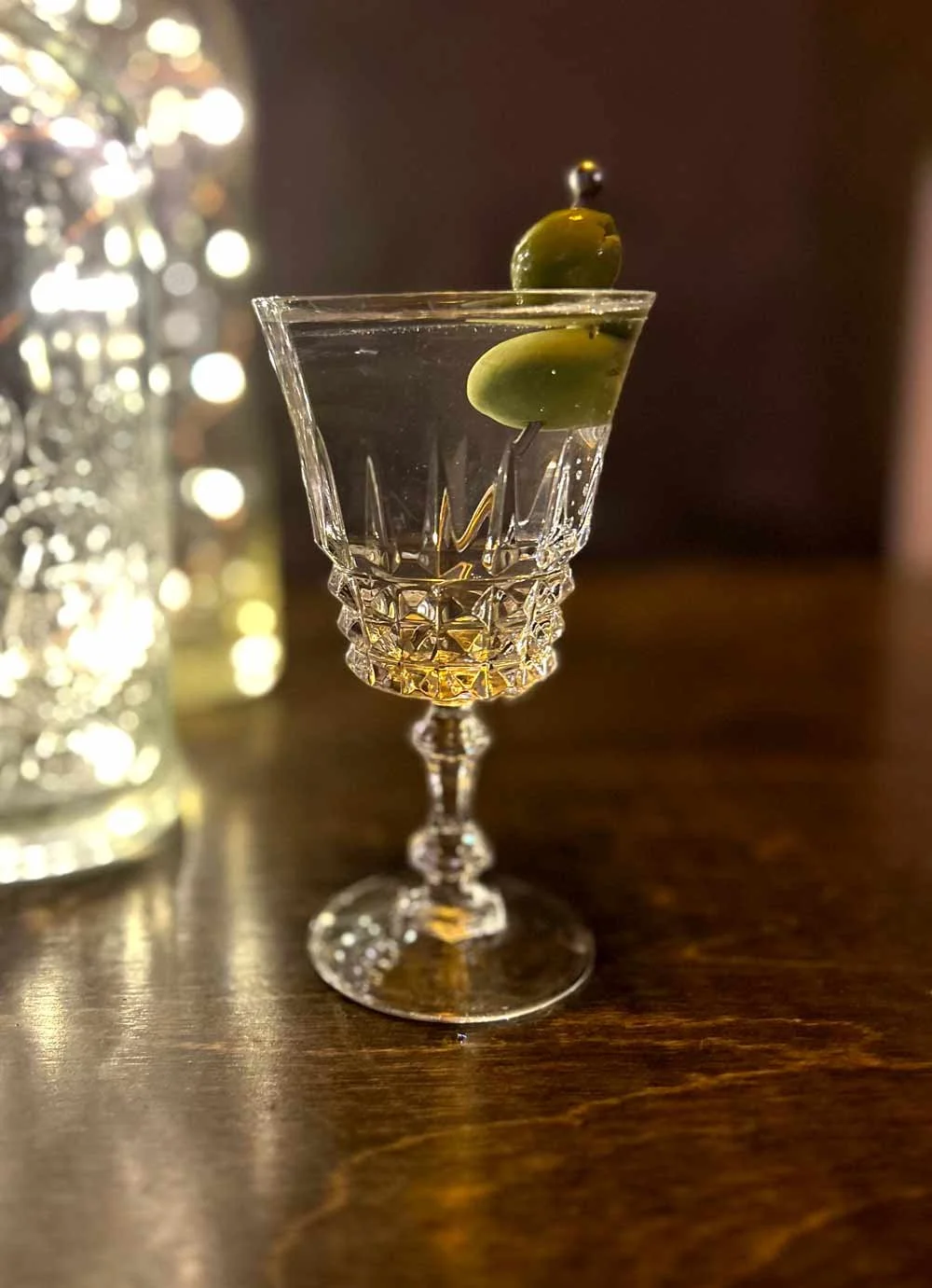 A crystal cocktail glass filled with a clear liquid, garnished with green olives, on a wooden surface with blurred decorative lights in the background.