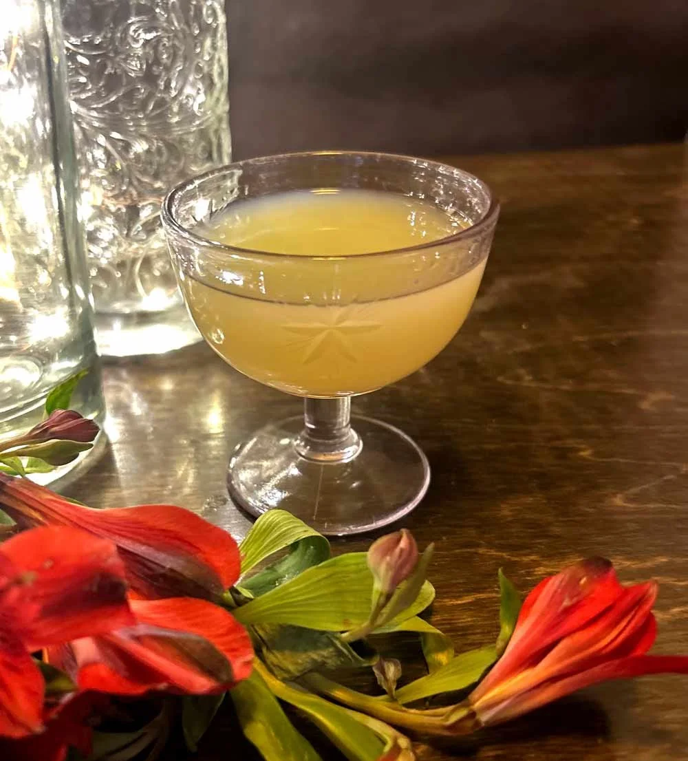 A glass of yellow cocktail on a wooden table, with a floral decoration and two clear textured glasses in the background.