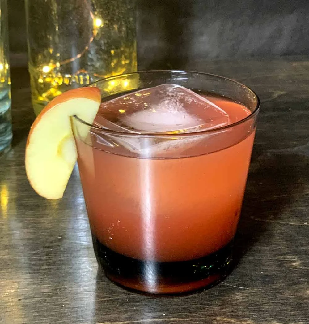 A pink cocktail with a lemon wedge garnish and a large ice cube in a short glass, on a dark wooden surface.