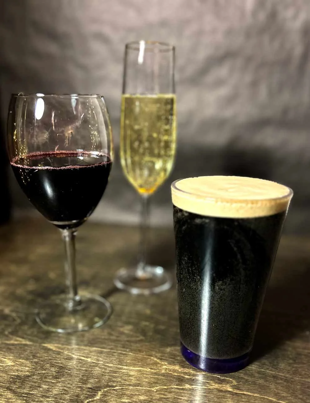Three glasses of beverages on a wooden surface, including a glass of red wine, a glass of sparkling wine, and a dark beer with a foam head.