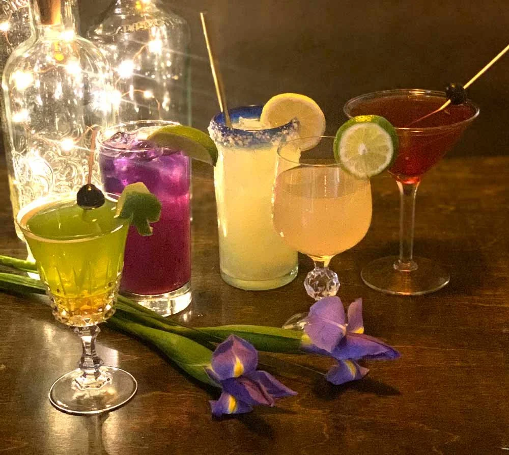 Five colorful cocktails with garnishes and a purple flower on a wooden table, illuminated by warm lighting.