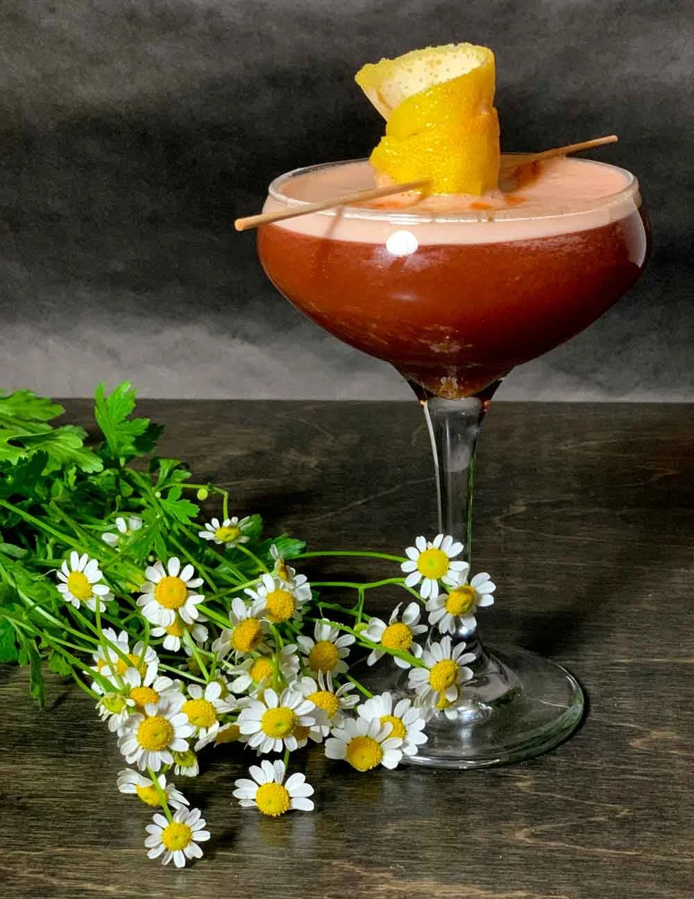 A pink cocktail in a coupe glass garnished with lemon and lime wedges on a wooden surface, accompanied by a bunch of white daisy flowers with yellow centers and green leaves, against a dark background.