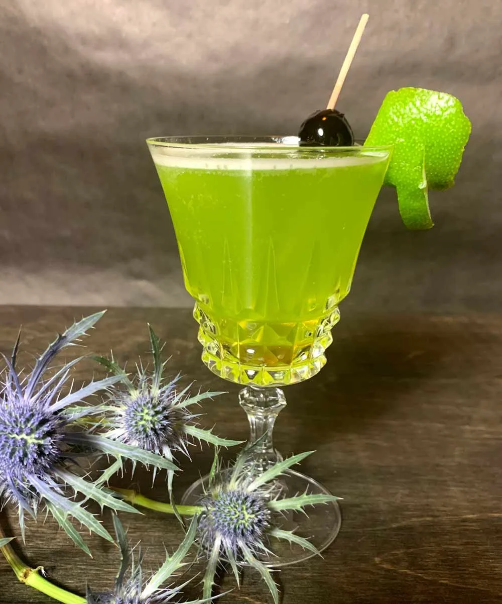 A green cocktail in a crystal glass garnished with a lime twist and a cherry on a stick, with some purple thistle flowers nearby.