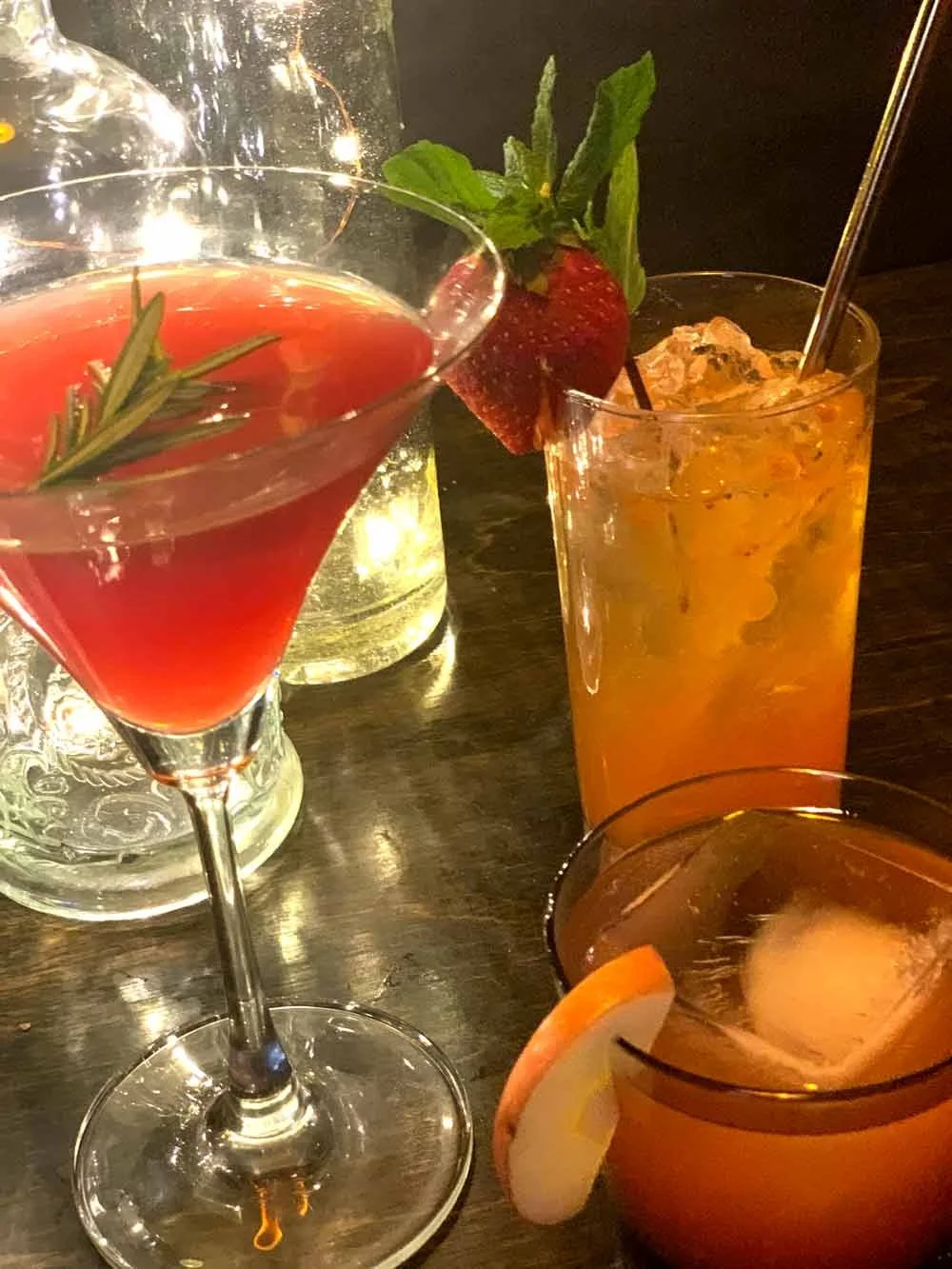 Three cocktails on a dark wooden table: a pink martini garnished with a sprig of rosemary and a strawberry, an orange-colored drink with ice and a straw, and an orange drink with a slice of orange and ice.