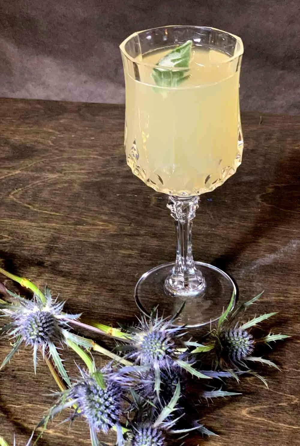 A glass of pale yellow cocktail garnished with a lime wedge, placed on a wooden surface with purple thistle flowers nearby.