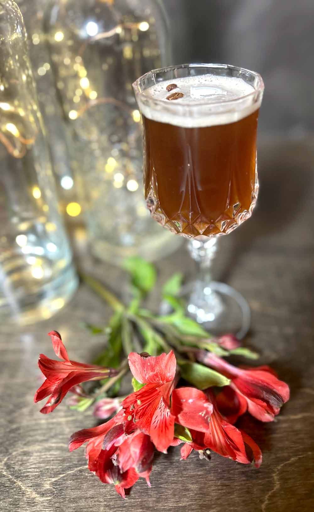 An espresso martini with coffee beans on top, blurred background with lights, pinkish-red flowers with green leaves on a wooden surface.