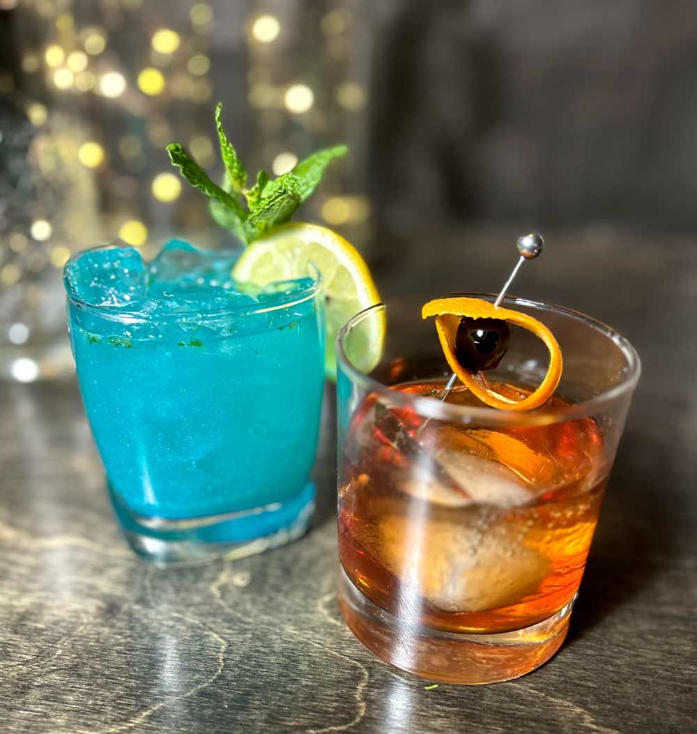 Two cocktails on a dark surface. One is a bright blue drink with ice, garnished with a lemon slice and a mint sprig. The other is an amber-colored drink with ice, garnished with an orange twist, a cherry, and a maraschino cherry on top.