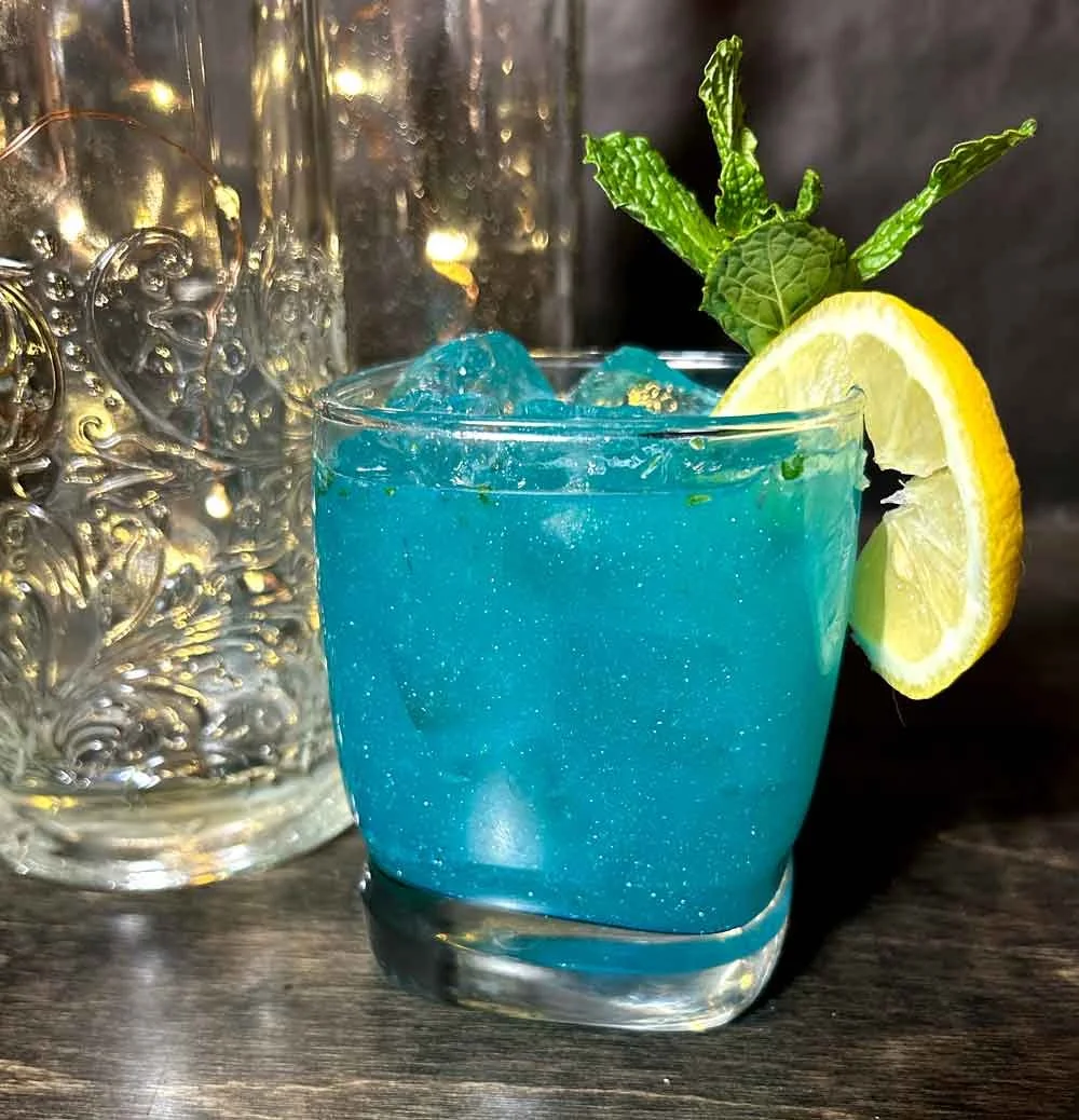 A glass filled with a blue cocktail, garnished with a lemon slice and fresh mint leaves, with ice cubes inside.