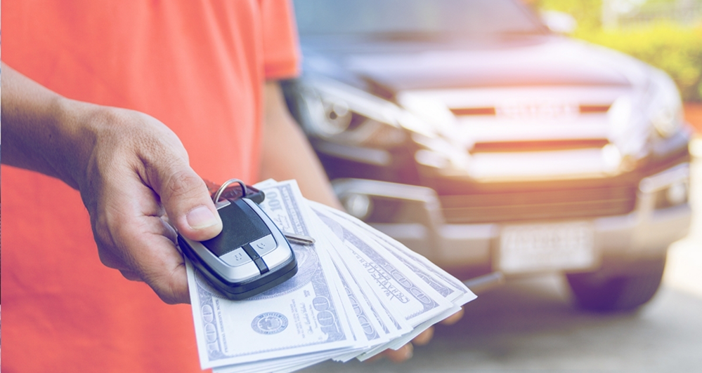 Person holding a car key and $100 bills in front of a black car.