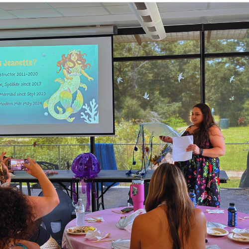 Young girl giving a presentation at a classroom or event, standing near a screen with a slide about a mermaid, with seated audience members taking photos, and large windows with trees outside.