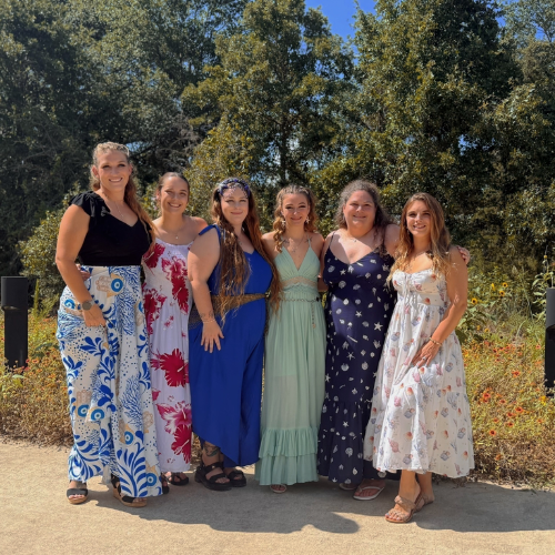Six women standing outdoors on a sunny day, dressed in colorful dresses, with trees and bushes in the background.
