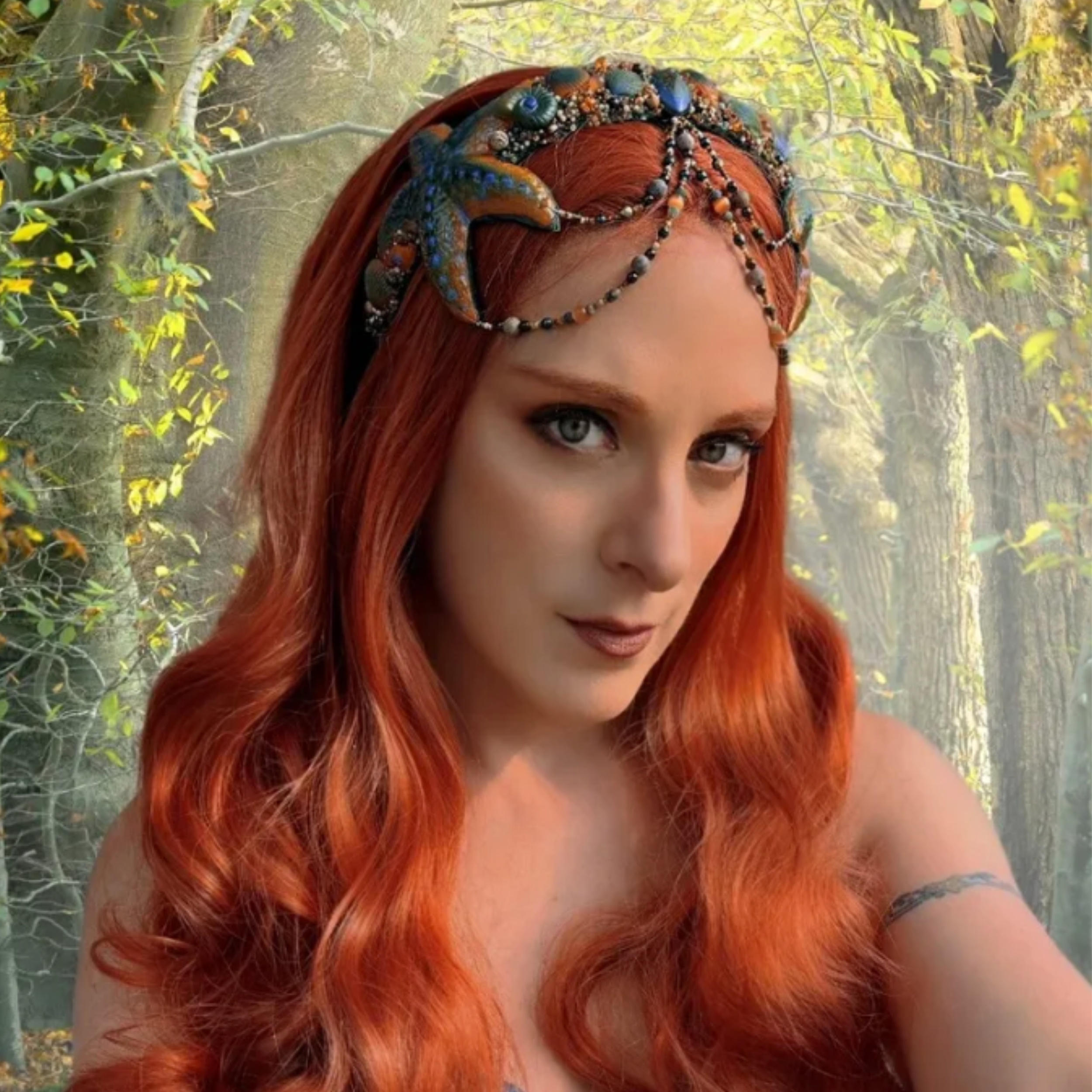 A woman with long, wavy red hair wearing a decorative headpiece with beads and sea creature motifs, standing in a forest with trees and foliage in the background.