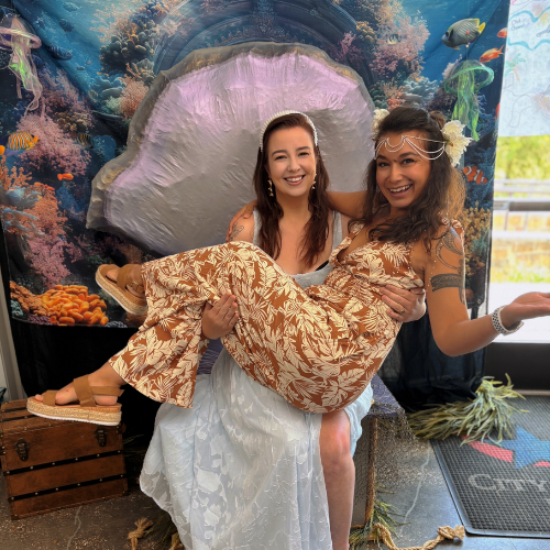 Two women smiling, one carrying the other, in front of an underwater-themed backdrop with coral and fish, with one wearing a floral dress and the other in a white dress, during a celebration or event.