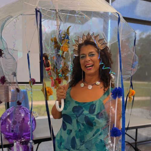 Woman wearing a festive crown and necklace, standing inside a colorful decorated tent with flowers and ribbons, smiling at the camera.