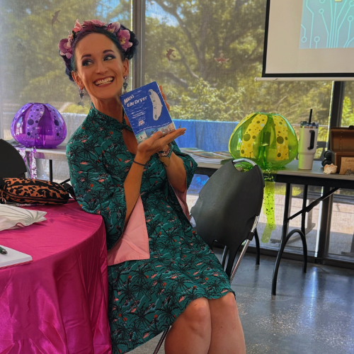 A woman with a floral headband sitting at a table, holding a box of MaxEbayer, smiling, with decorated lanterns and a presentation screen in the background.
