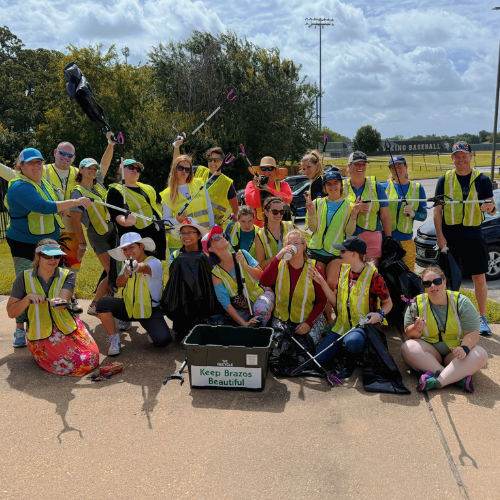 Group of people wearing yellow safety vests at a baseball field, holding litter pickers, with a sign that reads "Keep Brazos Beautiful."
