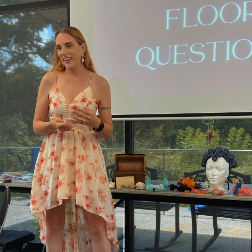 A woman in a floral dress stands in front of a presentation screen, gesturing as she speaks. There are various objects displayed on a table beside her, including a mannequin head with hair rollers, jewelry, and decorative items.