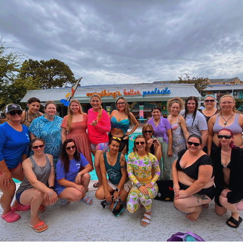 Group of women and girls posing in front of a colorful building with a sign that says "everything's better poolside". They are dressed in casual summer clothing, some are wearing sunglasses, and they are smiling.