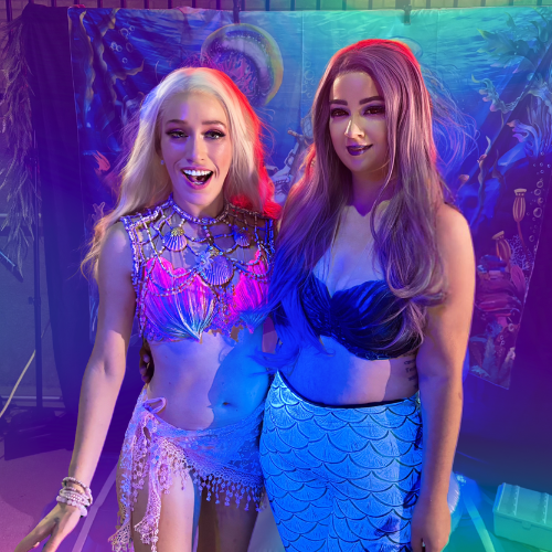 Two young women dressed as mermaids posing together at a costume event, with one wearing a silver and purple mermaid costume and the other a blue mermaid costume, under vibrant lighting.