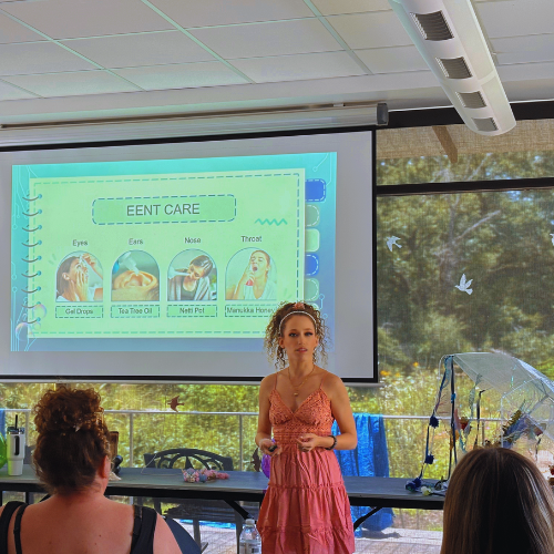 A woman in a pink dress giving a presentation on eye care to a small audience in a room with large windows showing trees outside.