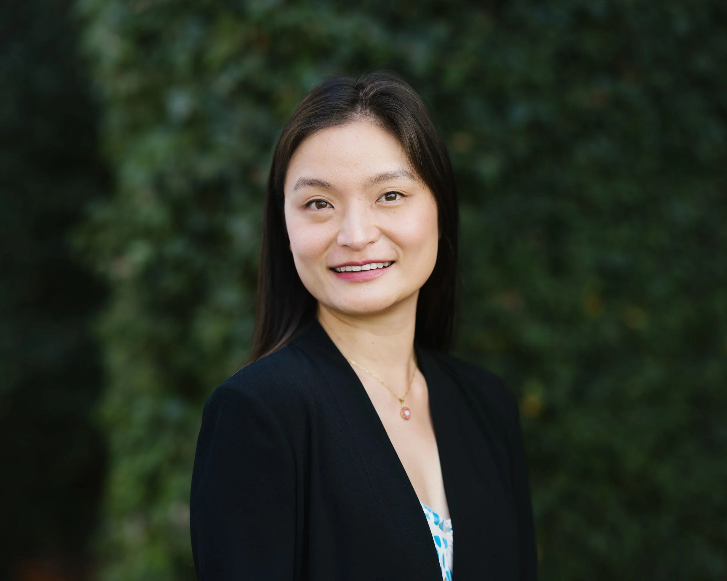 A woman with long dark hair smiling outdoors in front of greenery.