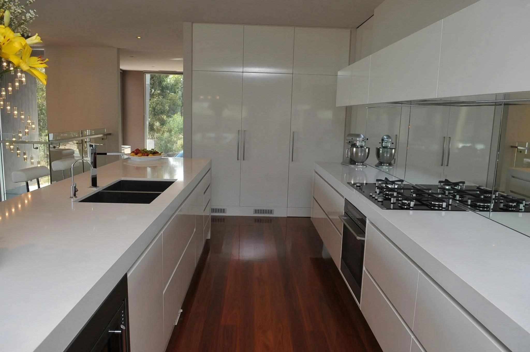 Modern white kitchen with a large island, black undermount sink, built-in oven, and a gas cooktop. There are two stand mixers on the countertop and a bowl of fruit near the window.