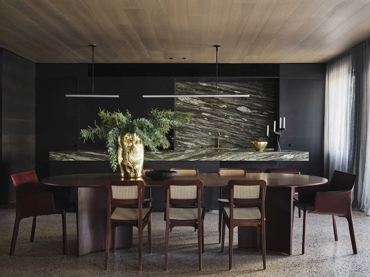 Modern dining room with a large oval wooden table, six chairs with wooden frames and beige cushions, a marble backsplash, a large gold vase with green plants, and a window with sheer curtains.