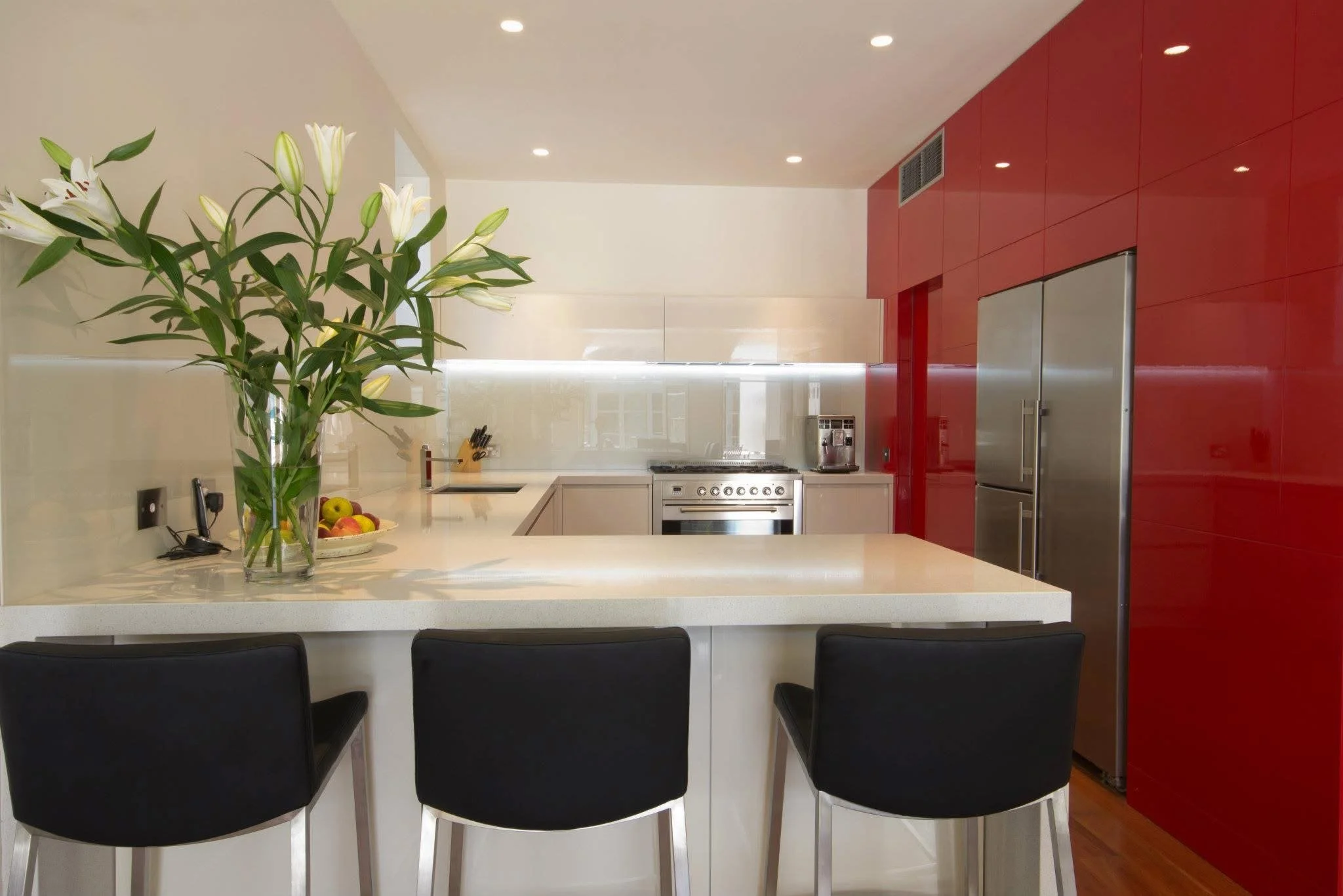 Modern kitchen with white countertops, red cabinets, stainless steel refrigerator, oven, and black chairs at the kitchen island, decorated with a large vase of white lilies and a bowl of apples.