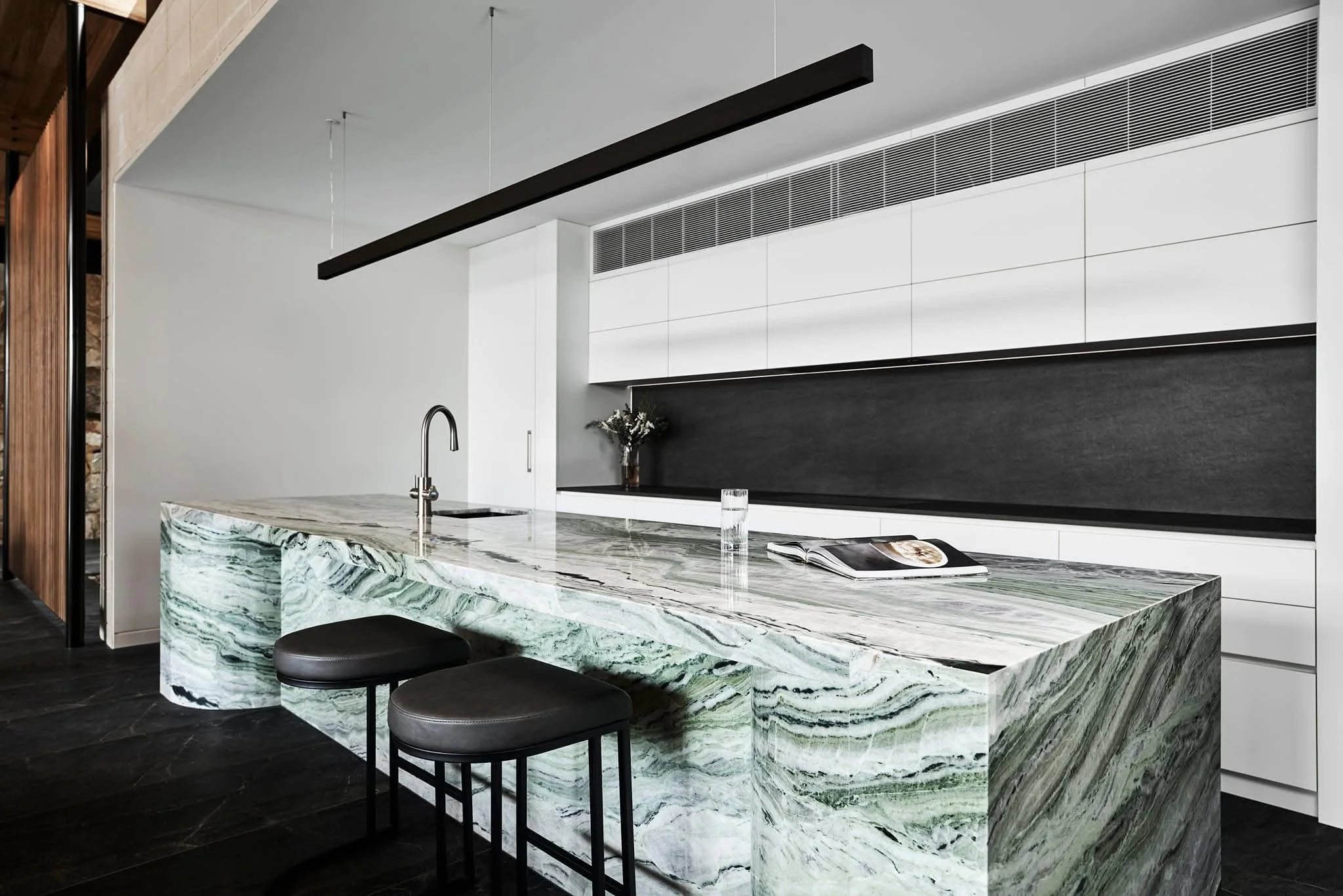 Modern kitchen with a marble island, two black stools, white cabinets, black backsplash, and a small bouquet of flowers on the counter.