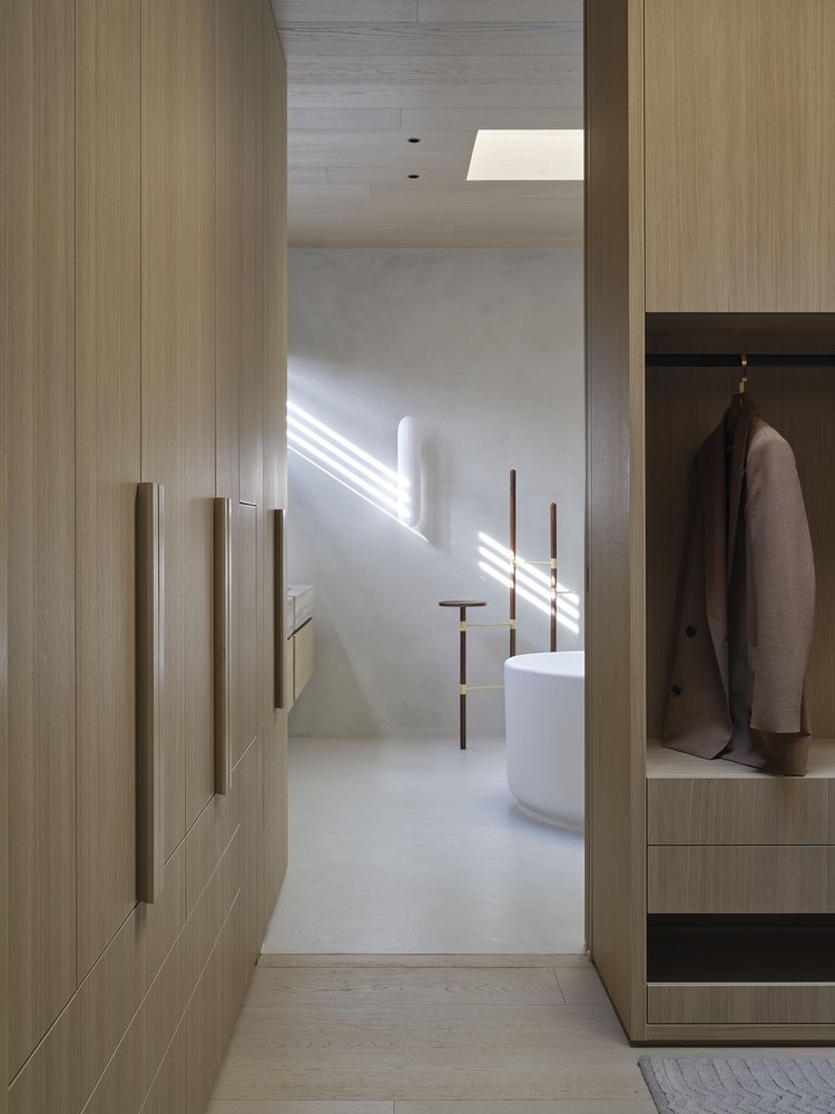A minimalist walk-in closet with wooden cabinets and drawers on both sides, a clothing hook with a blazer on the right, and modern decor including a white freestanding bathtub, two tall standing candlestick holders, and natural light streaming through a rectangular skylight and attached window creating diagonal light patterns on the wall.