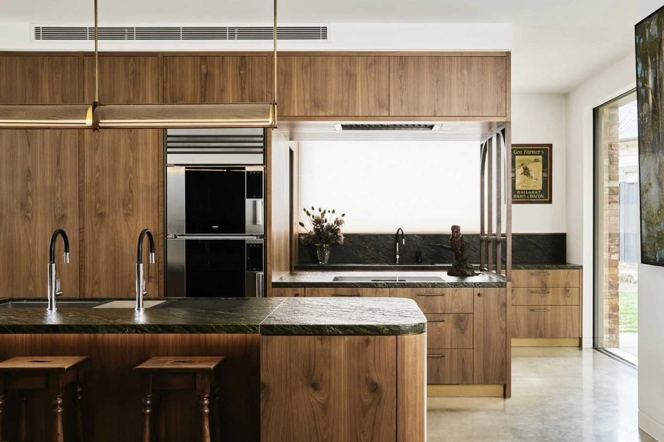 Modern kitchen with wooden cabinets, black countertops, a built-in oven, and an island with two stools. There are two black faucets, a vase with flowers, and a framed vintage poster on the wall.