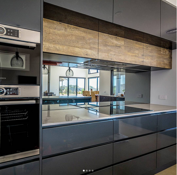 Modern kitchen with dark gray cabinetry, a light-colored countertop, and wooden upper cabinets. Reflections on the countertop show a dining area with large windows and a view outside.