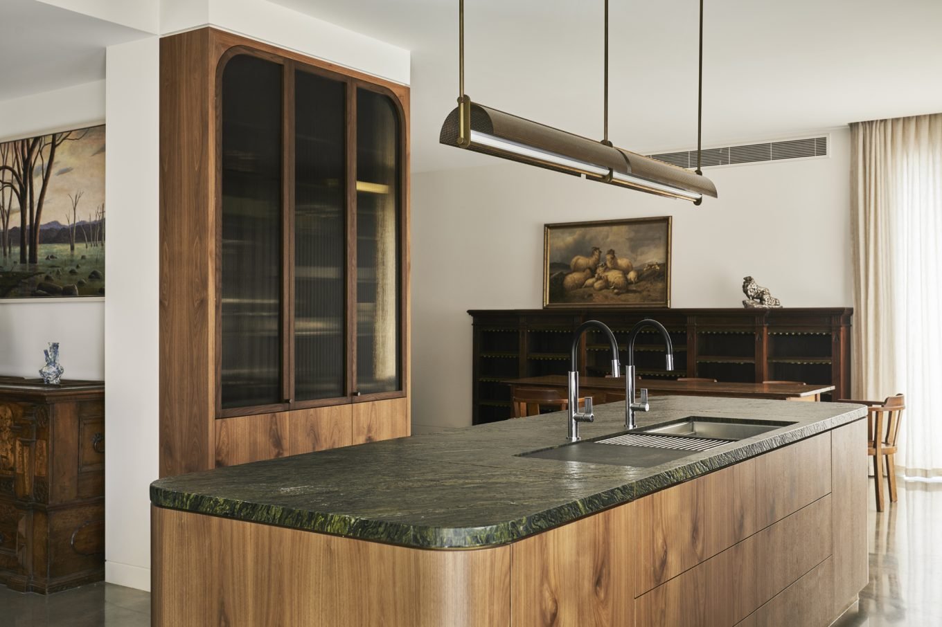 Modern kitchen with a wooden island, dark green marble countertop, stainless steel sink, wooden cabinet, and a dining area with a dark wood shelf, a painting of sheep, and a decorative object, illuminated by natural light from curtains.
