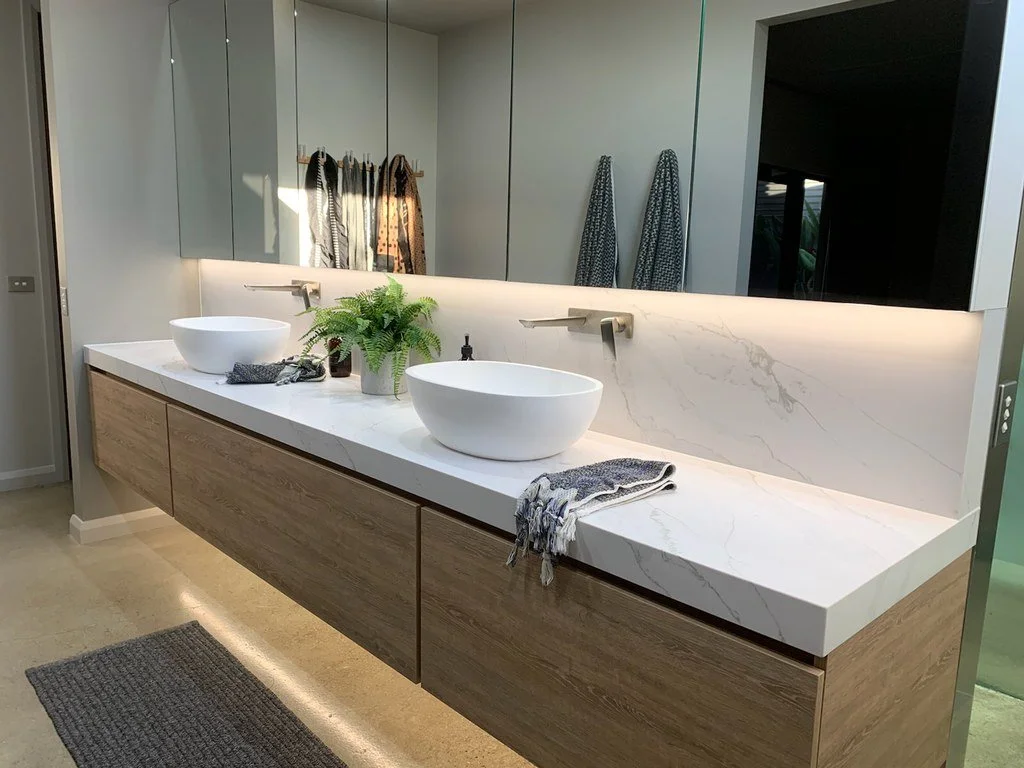Modern bathroom vanity with two vessel sinks, a large mirror, and wooden cabinets, decorated with a green plant and towels.