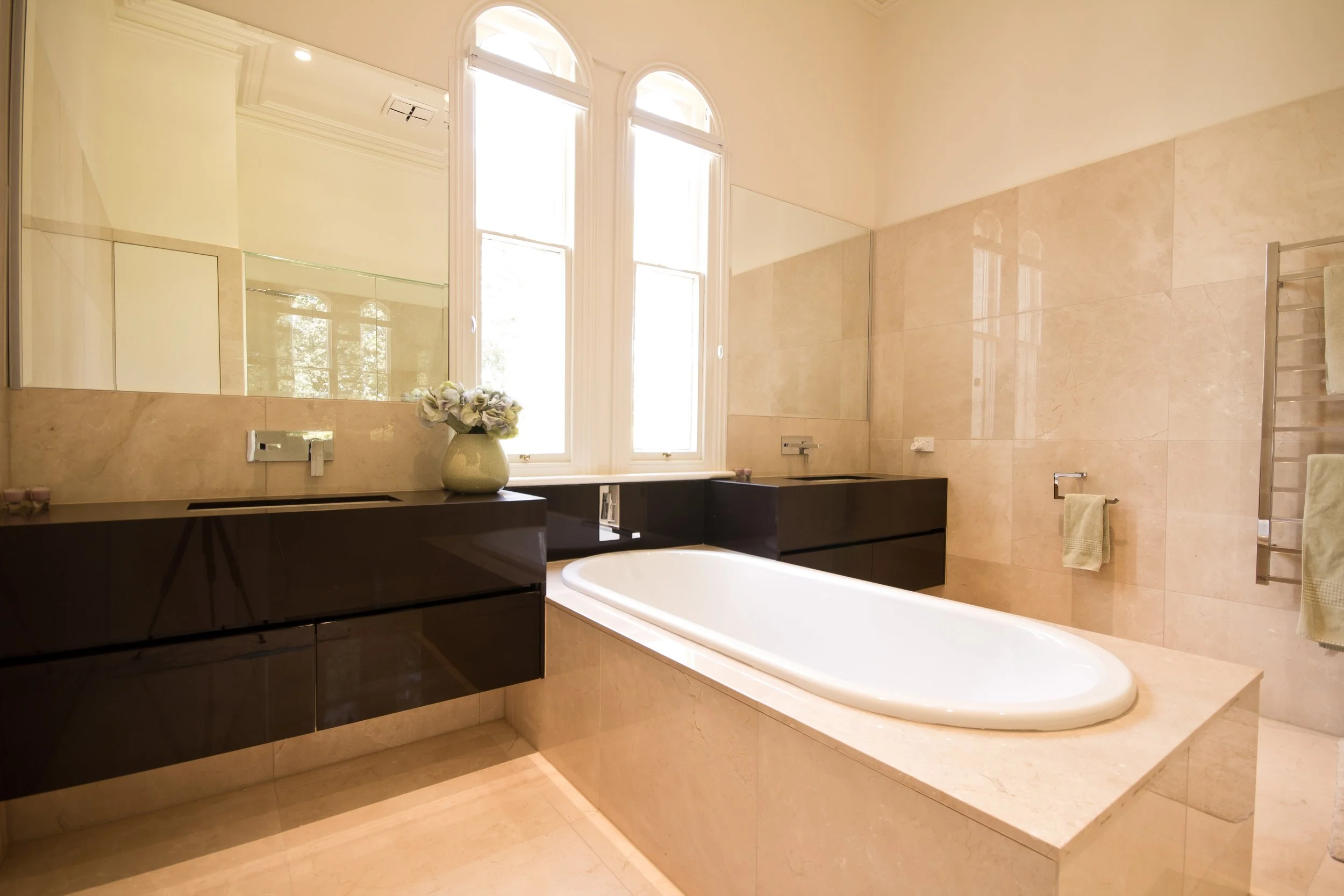 Bathroom with a large window, bathtub, black vanity, flower vase, towel rack, and beige tiled walls and floor.