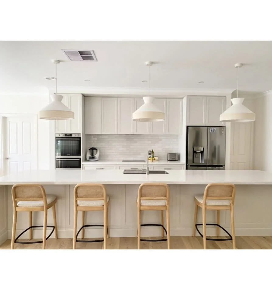 Modern kitchen with white cabinets, a large white island, four beige barstools, pendant lighting, stainless steel appliances including a refrigerator, oven, and microwave, and minimalist decor.