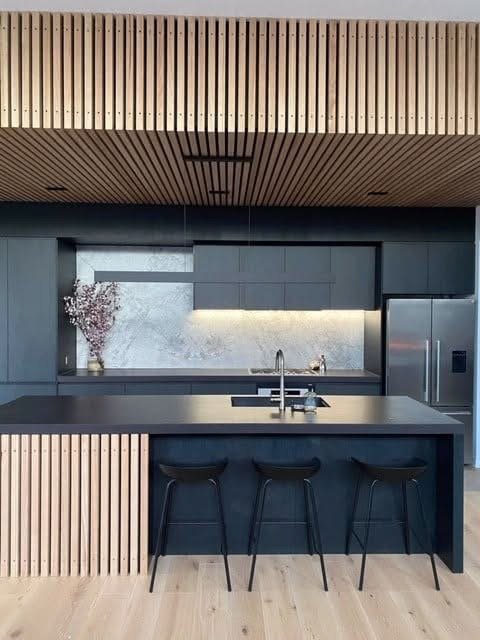 Modern kitchen with black cabinetry, wooden ceiling with linear panels, gray backsplash, stainless steel appliances, and a black island with three bar stools.