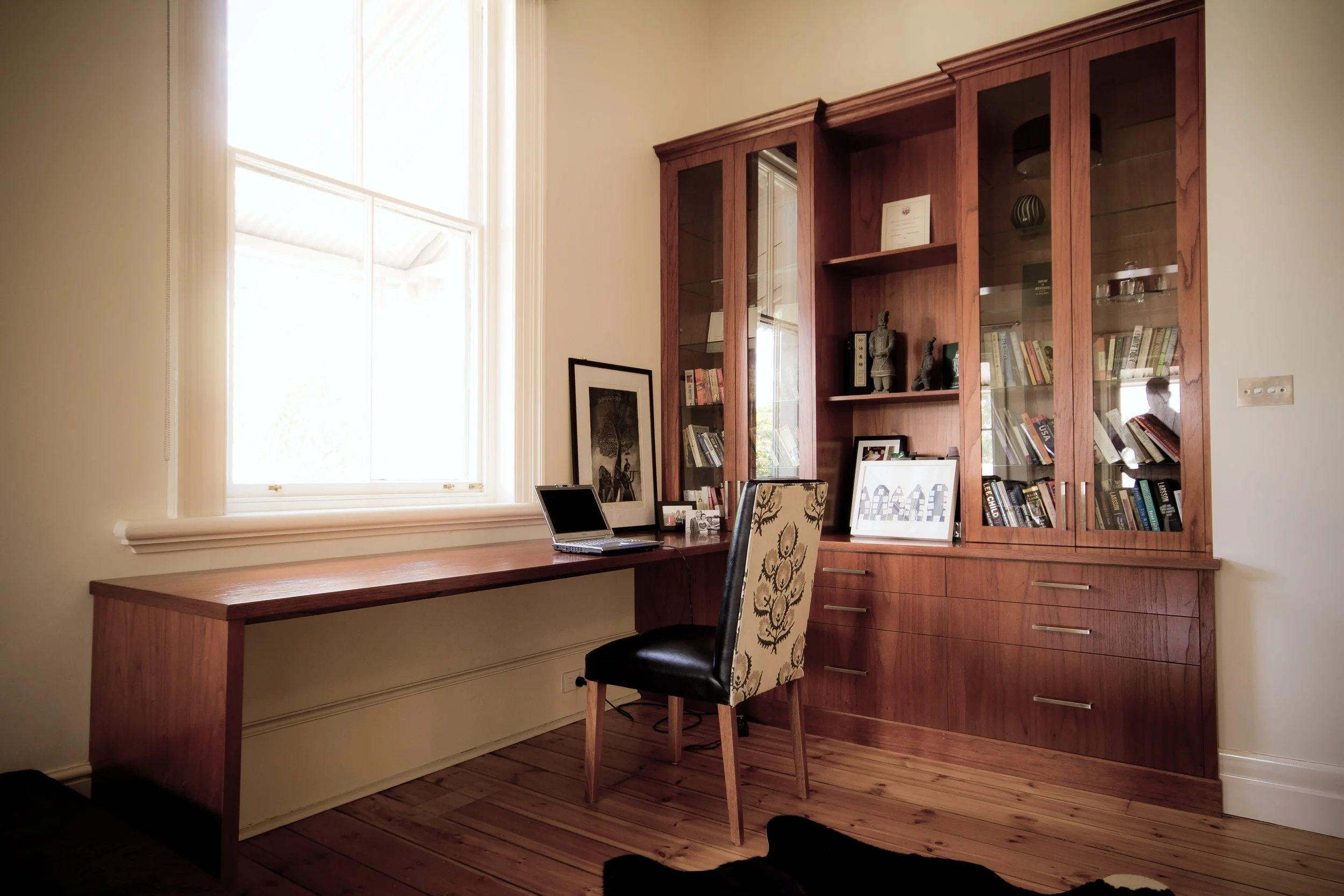 Home office with wooden desk, black and patterned chair, glass-fronted bookshelf filled with books and decorations, large window with white trim, framed artwork, and hardwood flooring.