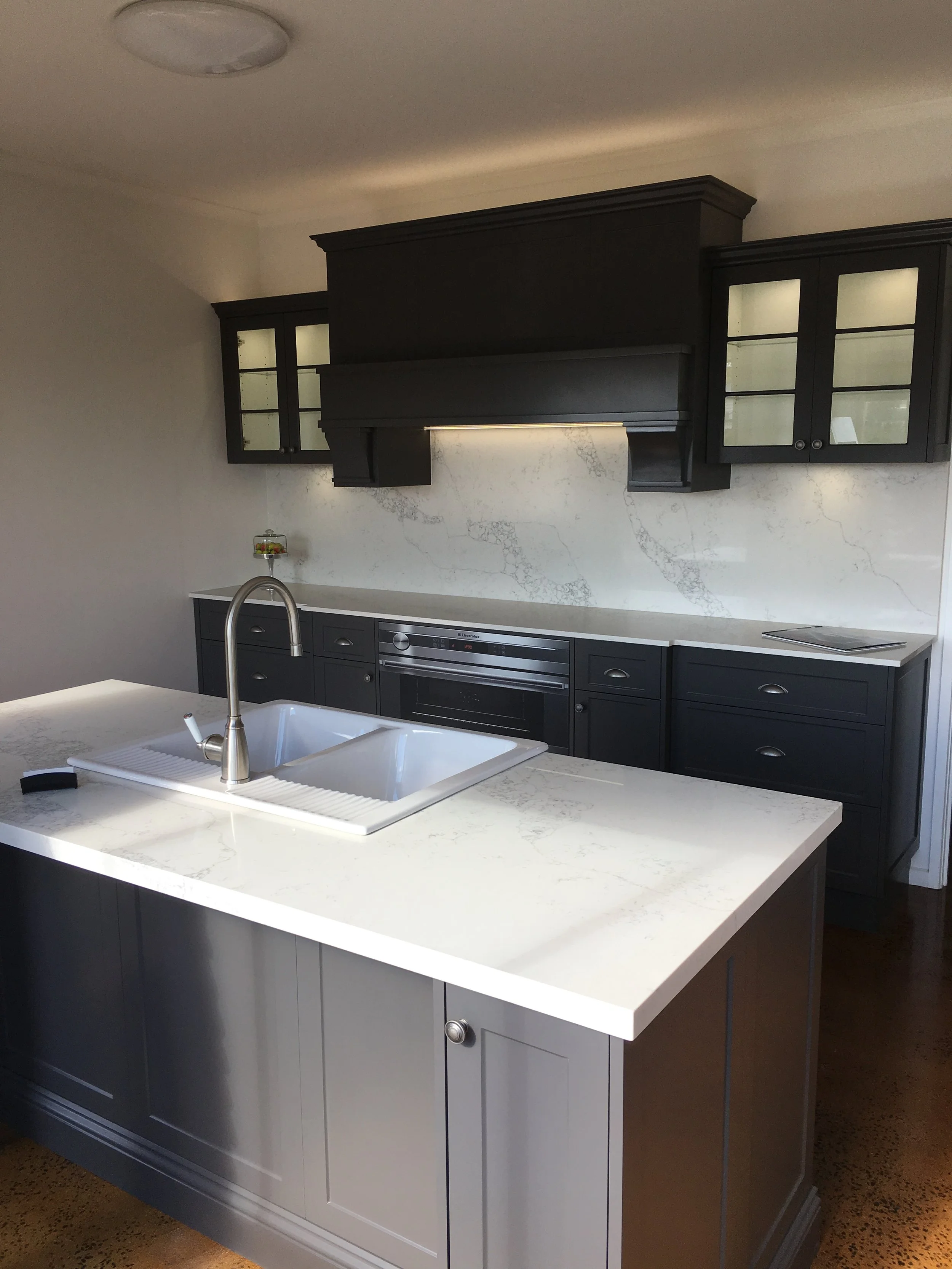 Modern kitchen with white marble countertop island, black cabinets, and a white backsplash with veining. There is a double-basin sink with a tall faucet and a stovetop on the counter. Upper cabinets have glass-pane doors, and the kitchen has a ceiling light fixture.