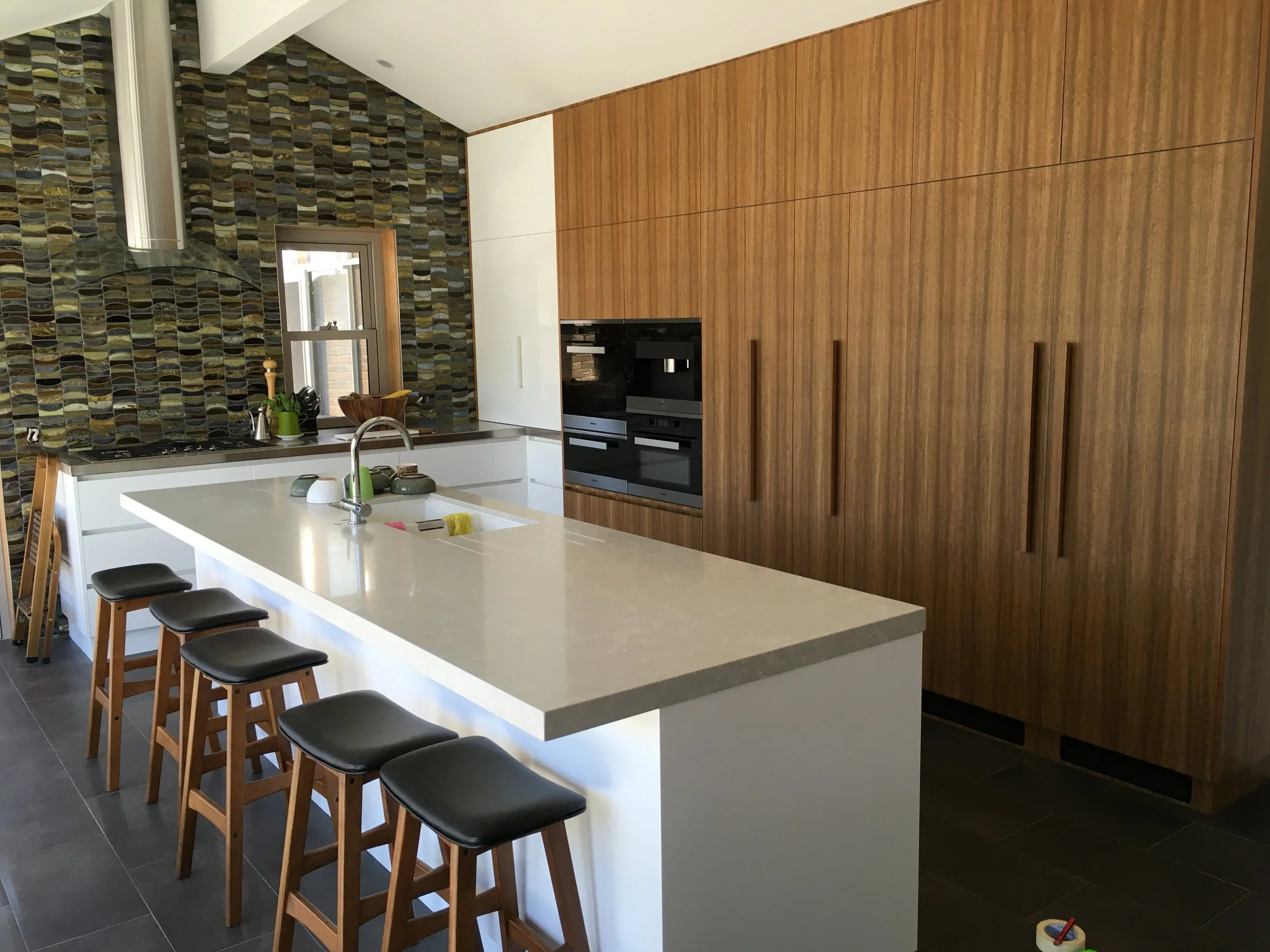 Modern kitchen with white island, black stools, wood cabinets, tiled wall, and small window