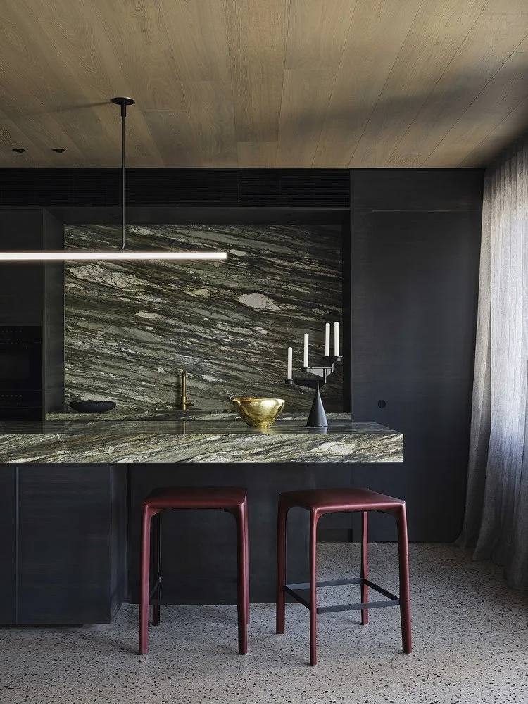 Modern kitchen with a marble countertop and backsplash, two red bar stools, black candlestick holder with white candles, gold bowl, and a black bowl, with beige terrazzo flooring and a wood ceiling.
