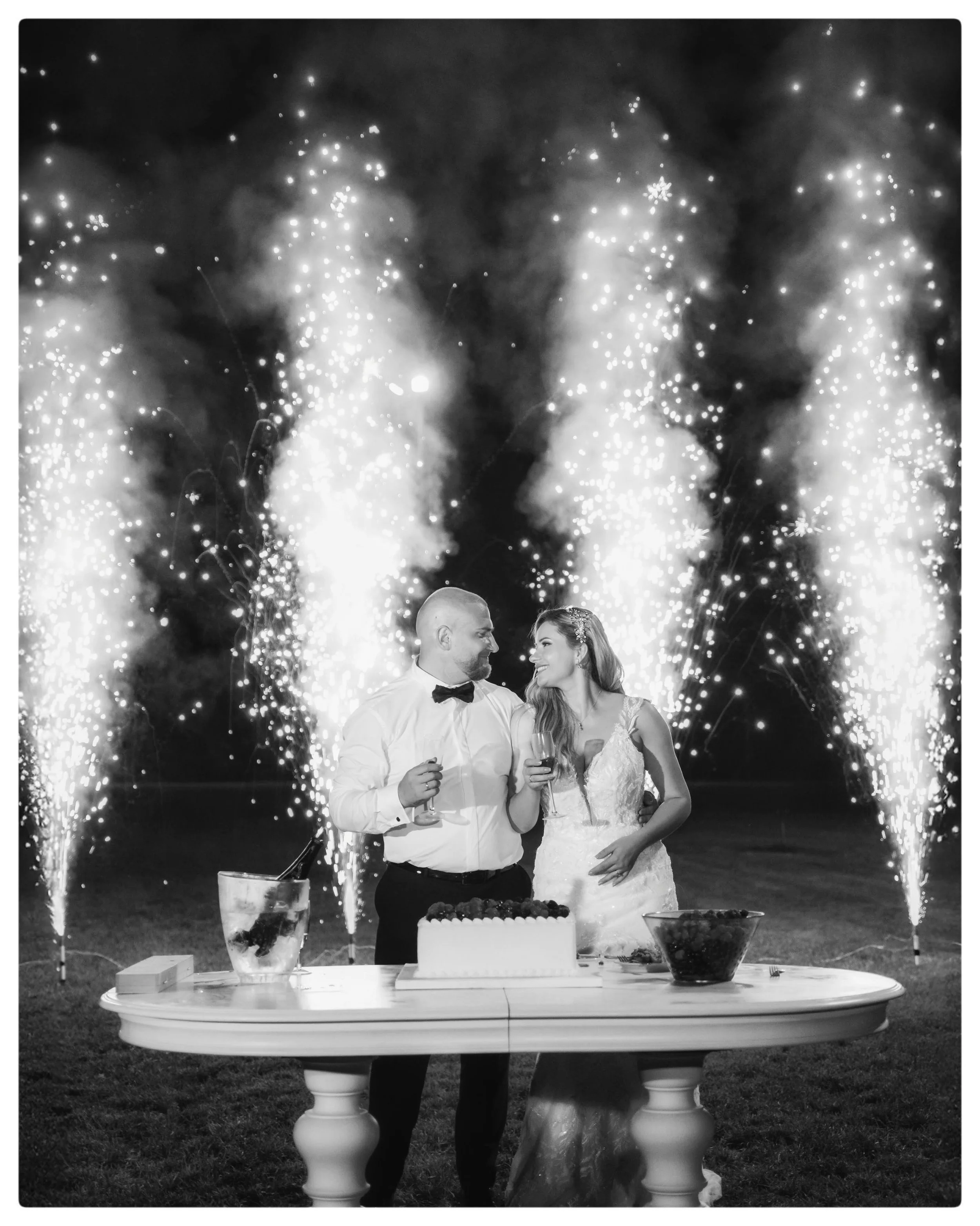 A black-and-white photograph of a newlywed couple standing under a display of fireworks, smiling and holding wine glasses, while a wedding cake and desserts are on the table in front of them.