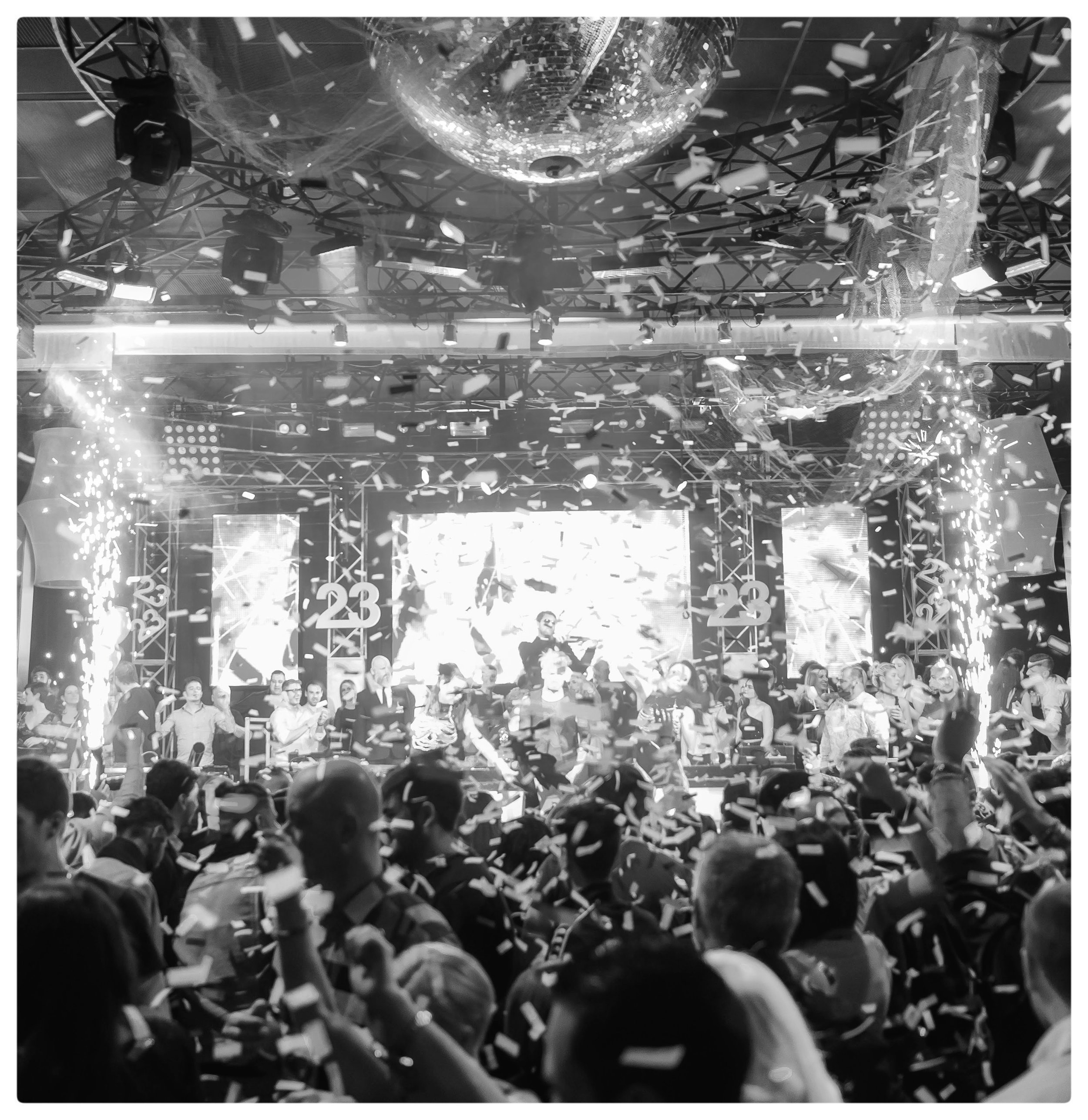 People attending a celebration or event with a large disco ball hanging from the ceiling, confetti falling, bright stage lights, and a crowd enjoying the occasion.