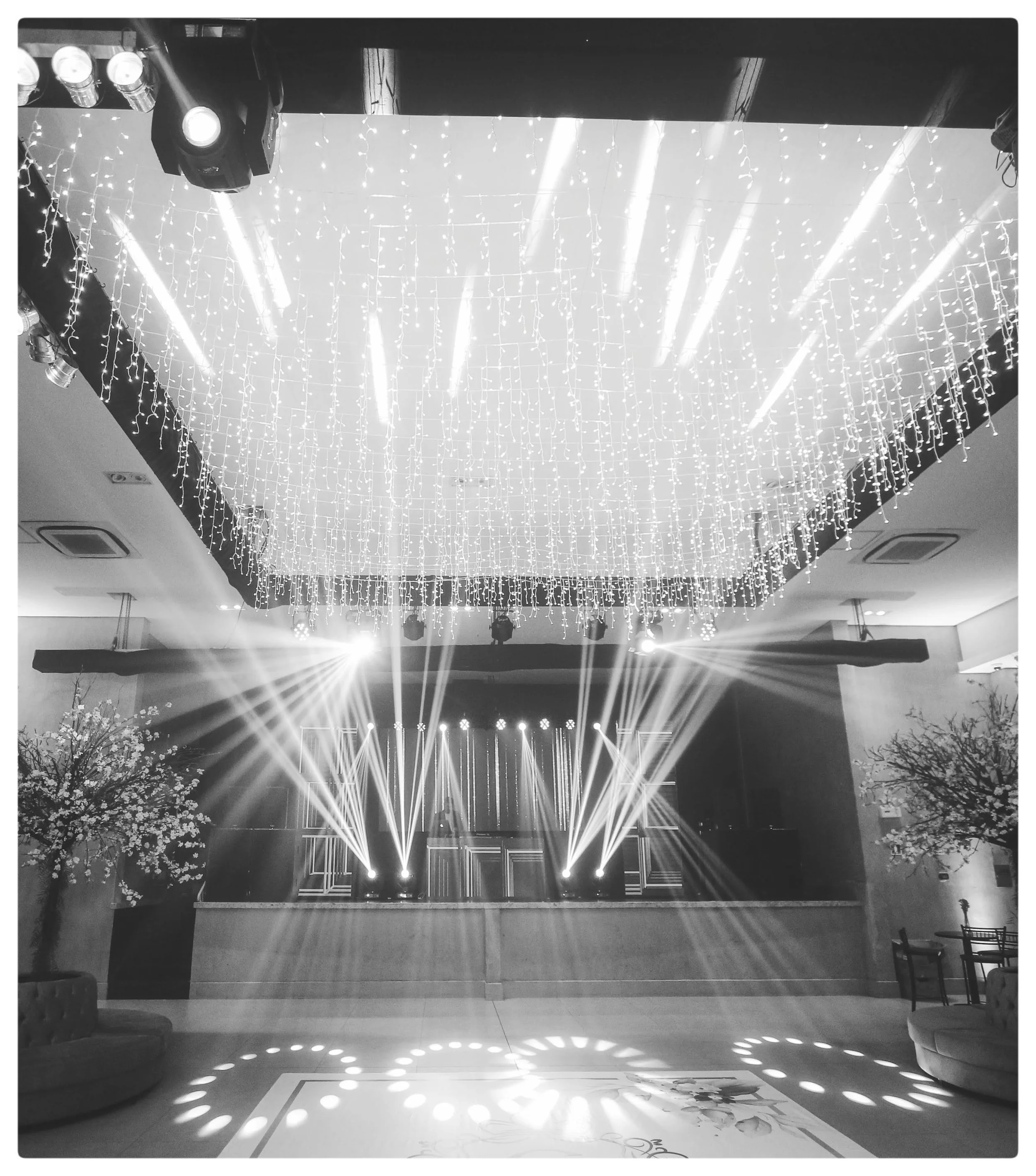 Black and white photo of a decorated indoor event space with hanging string lights, stage lighting, and a stage with geometric light designs, floral arrangements, and seating area.