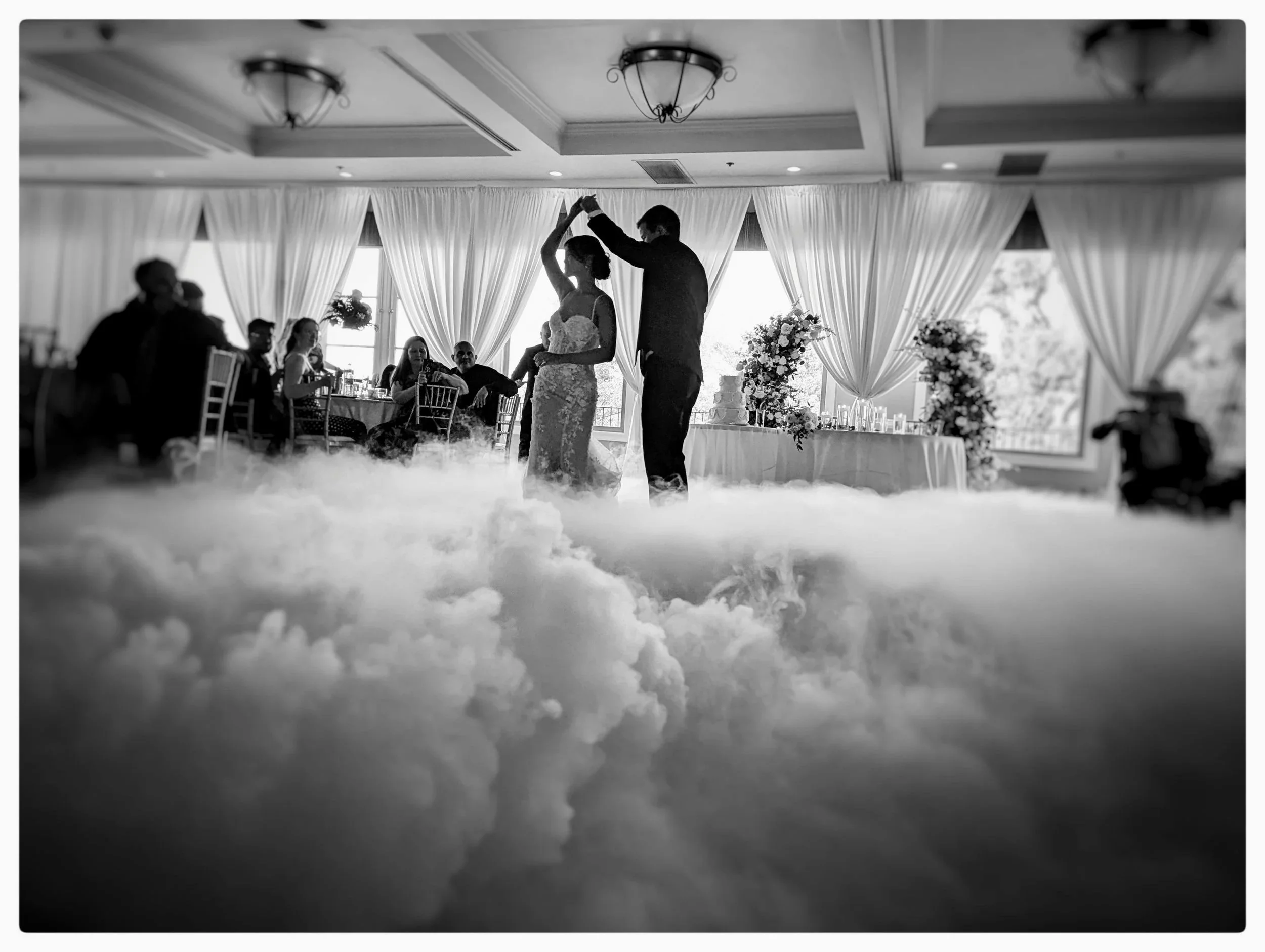 A black and white photo of a bride and groom dancing on a cloud-like layer of fog at their wedding reception, with guests seated at tables in the background and large windows with curtains behind them.