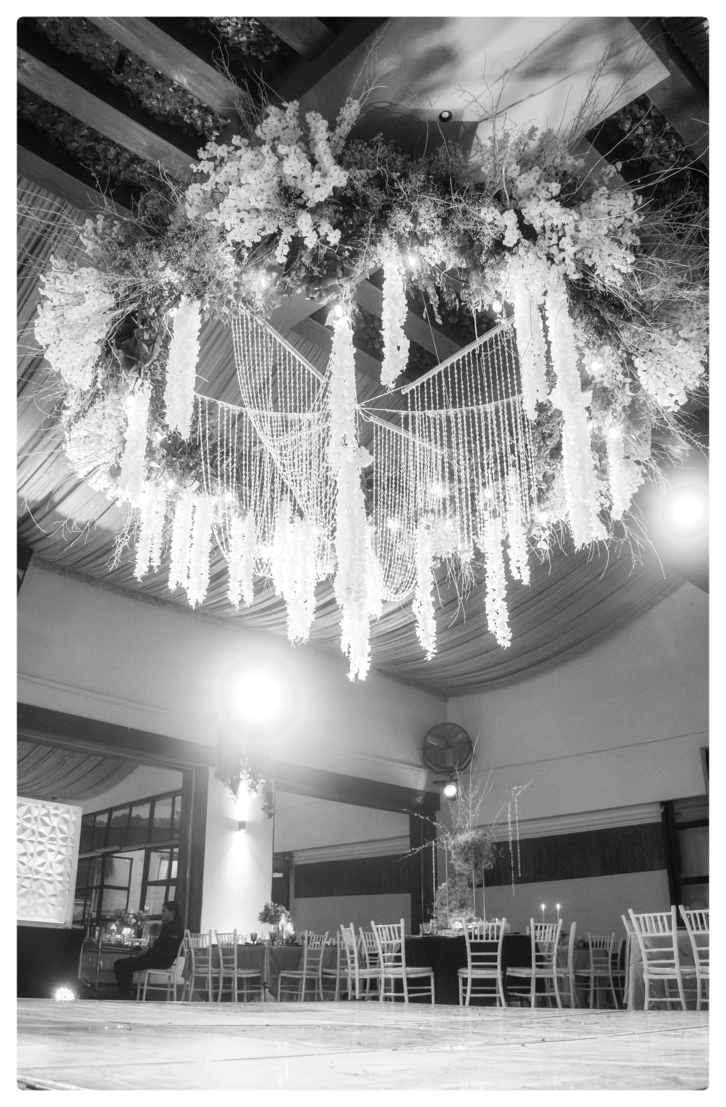 Elegant venue with a large floral chandelier hanging from the ceiling, wooden floor, round tables with chairs, and a person sitting at one table. The decor suggests a formal event or celebration.